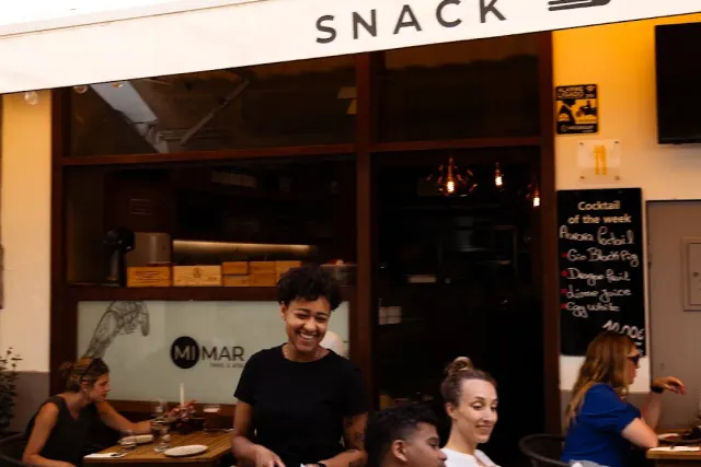 A smiling staff member at an open front with a cocktail board and diners outside