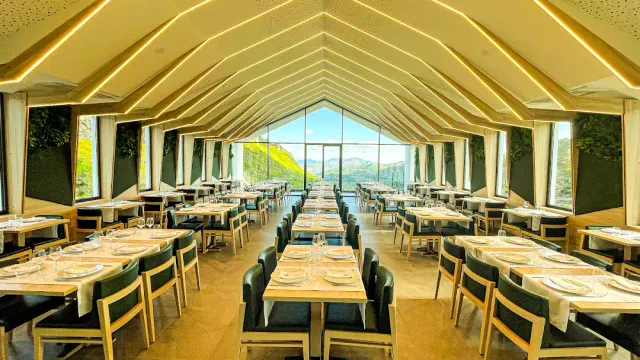 A long room with a lit pitched ceiling and set tables facing a window onto mountains
