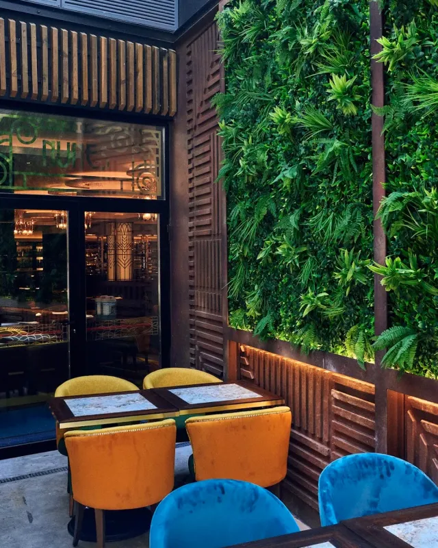 A courtyard with a green plant wall, orange and blue velvet chairs and small tables