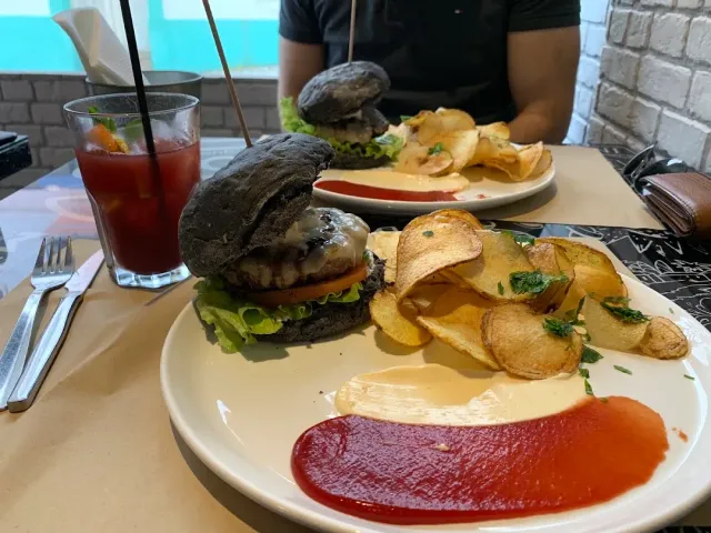 Two plates with a burger in a black bun, homemade crisps and sauce, and a glass of a red drink