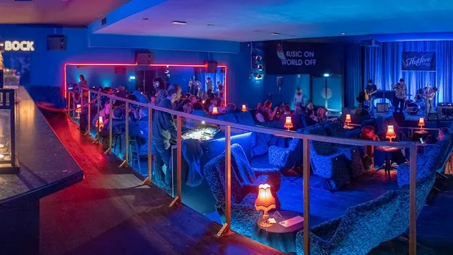 A blue-lit hall with armchairs, table lamps and a band playing on the stage