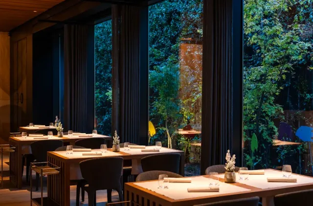 A dark room with set tables and tall windows onto a lit garden