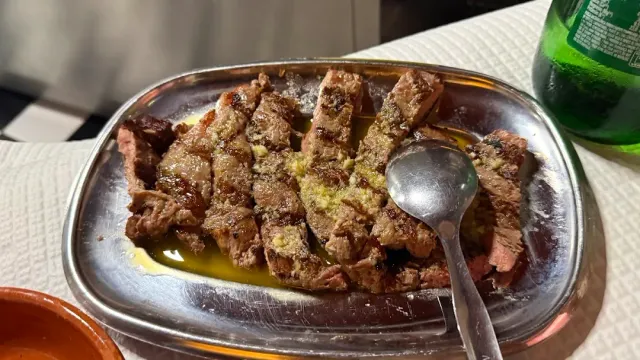 A metal tray with strips of roasted meat in oil and a spoon laid beside them
