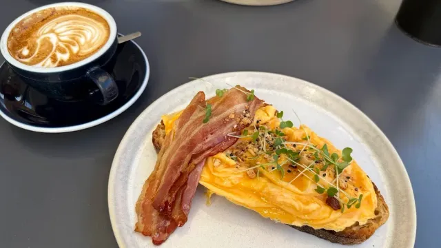 A slice of bread with scrambled eggs, a strip of bacon and sprouts, beside a cappuccino in a dark cup