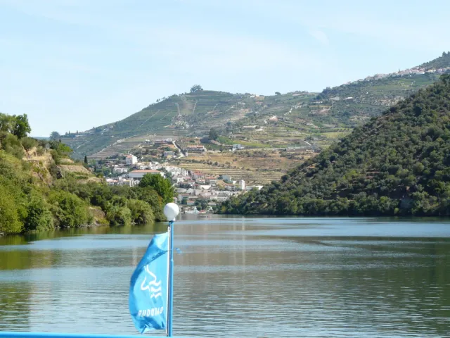 A boat bow with a light blue flag facing a wide river and a village on a terraced slope
