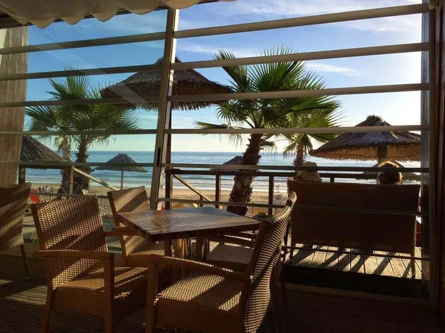 Wicker chairs and a table on a wooden terrace facing palms, straw umbrellas and the sea