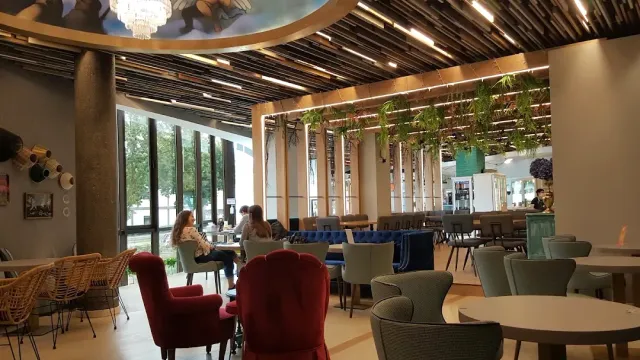 A spacious seating hall with hanging plants, red armchairs and round tables