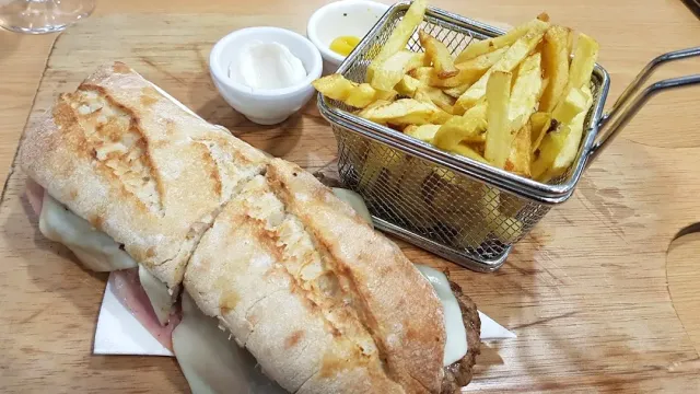 A sandwich in rustic bread cut in half beside a basket of fries and two bowls of sauce