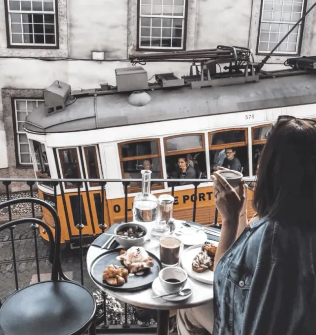 A woman eating on a balcony above a yellow tram, a table with pastries and coffee cups