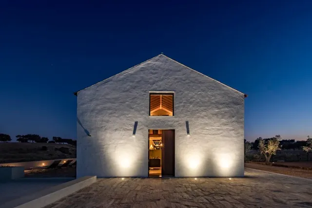 A whitewashed building in the evening with a lit doorway and a stone forecourt