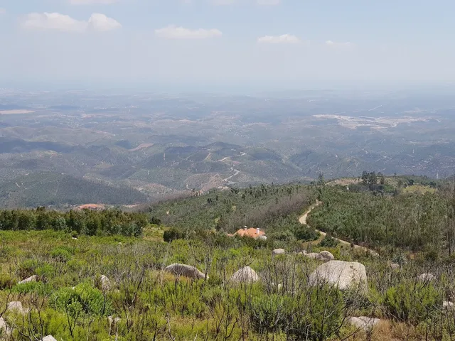 A wide view from a rocky summit over forested ridges and a plain stretching to the horizon
