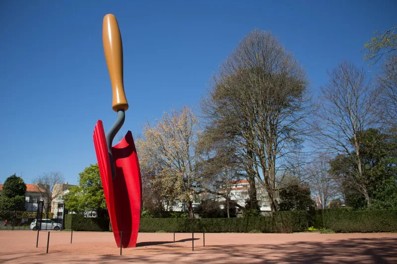 A giant red trowel sculpture on a lawn among trees under blue sky