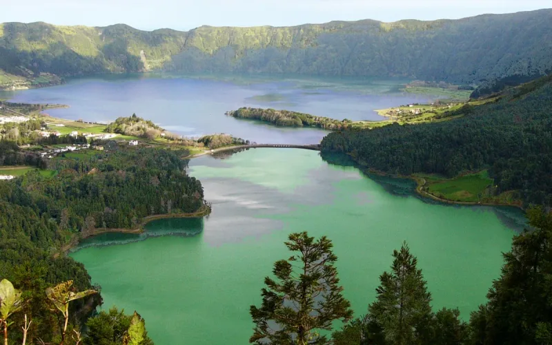 Two adjoining crater lakes, one blue and one green, separated by a bridge among wooded hills