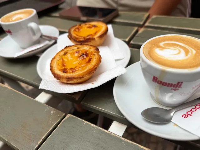 Two golden custard tarts and cups of coffee with foam on an outdoor wooden table