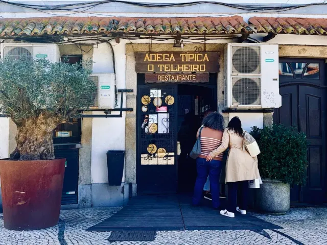 An entrance with a yellow sign and a tiled roof, two women in the doorway and an olive tree in a pot