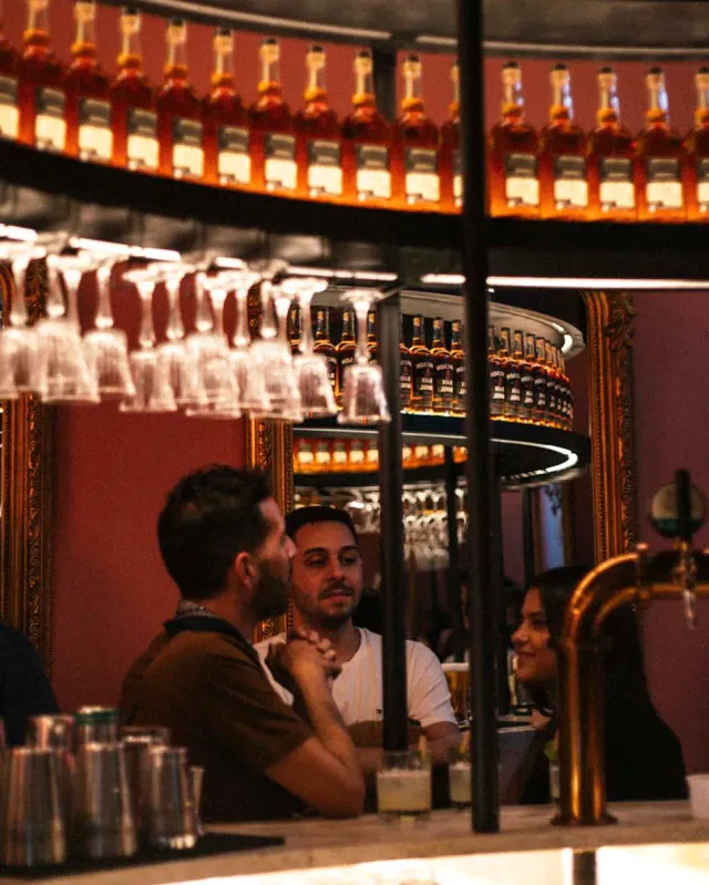 Lit rows of bottles above a counter, hanging glasses and guests chatting