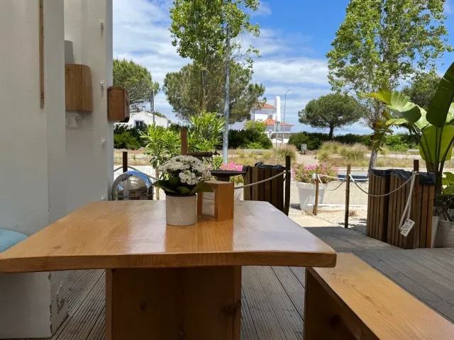 A wooden table and bench on a deck, a pot of white flowers, pines and a white house behind