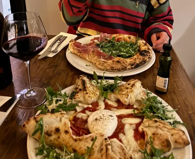 Two pizzas on a wooden table with a glass of red wine and a bottle of olive oil