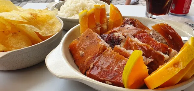 Pieces of roasted pork with crisp skin beside orange slices, rice and fried potatoes