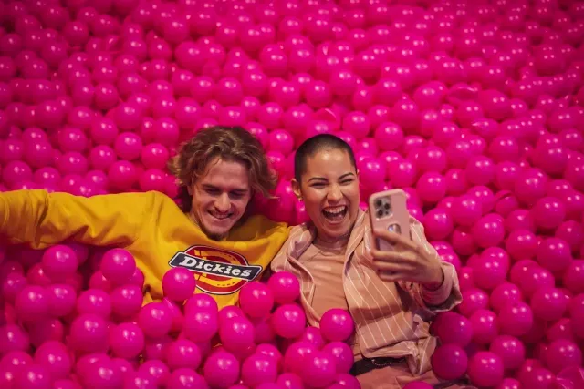 A laughing couple sinking into a pink ball pit and taking a photo of themselves