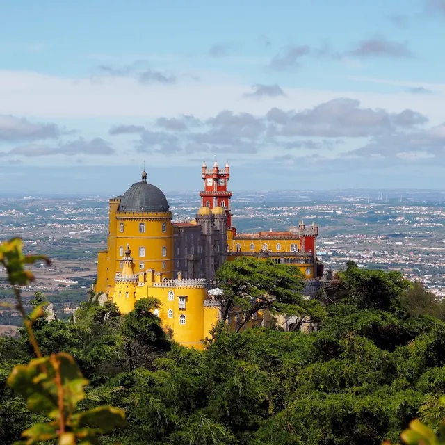 A yellow and red palace with a dome and a clock tower above a green forest and a wide plain