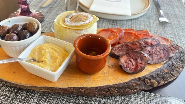 A wooden board with olives, cheese, sausage slices, a clay pot and a yellow spread