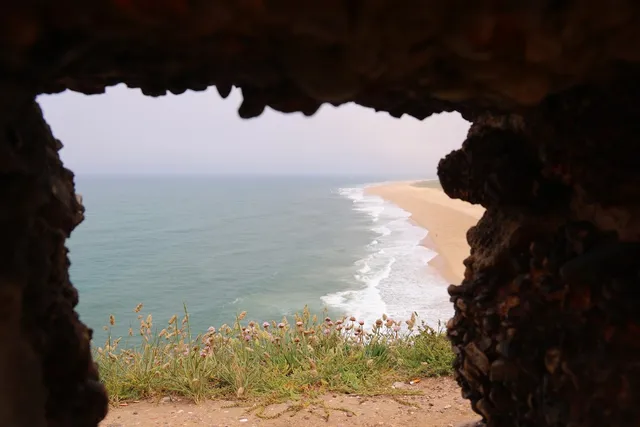 An opening in a stone fortress wall, with a long sandy beach and white waves beyond