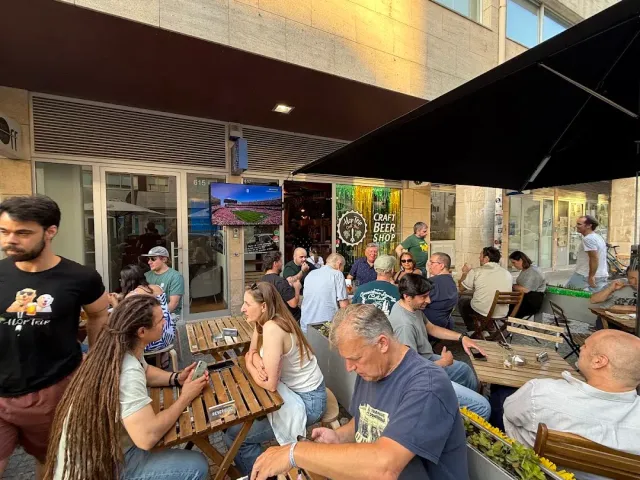 A busy forecourt with people sitting on wooden benches outside a beer shop