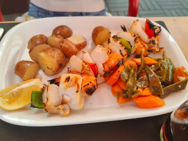 A plate of grilled squid skewers, small potatoes, sautéed vegetables and a lemon wedge