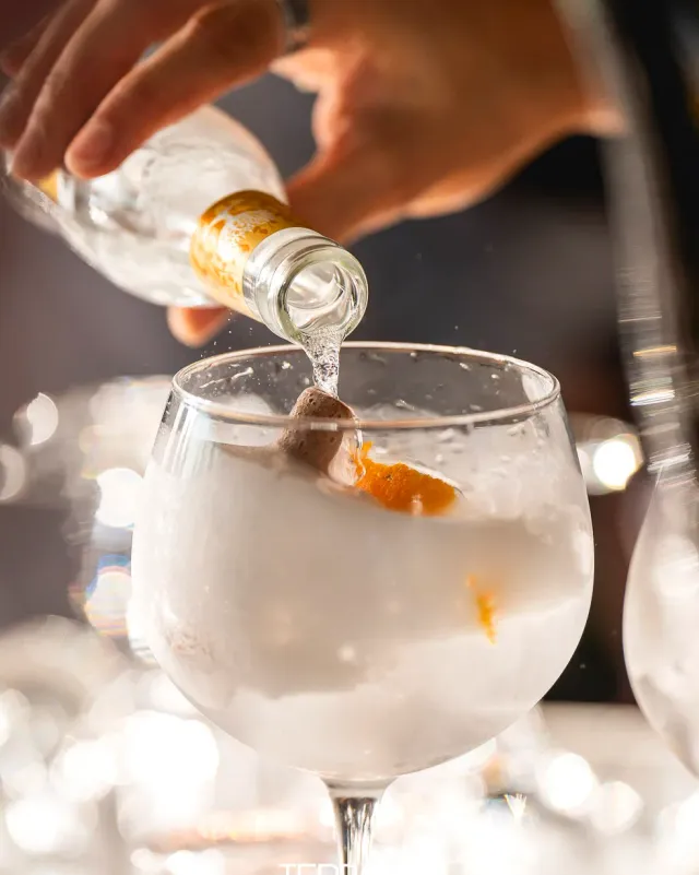A hand pouring tonic into a goblet of ice with orange peel, blurred lights behind