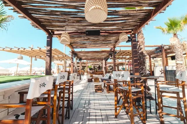 A wooden deck under a reed pergola with rattan lamps and decorated chairs