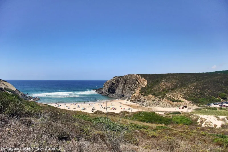 A sandy cove with a headland jutting into blue sea, seen from a scrub covered hill