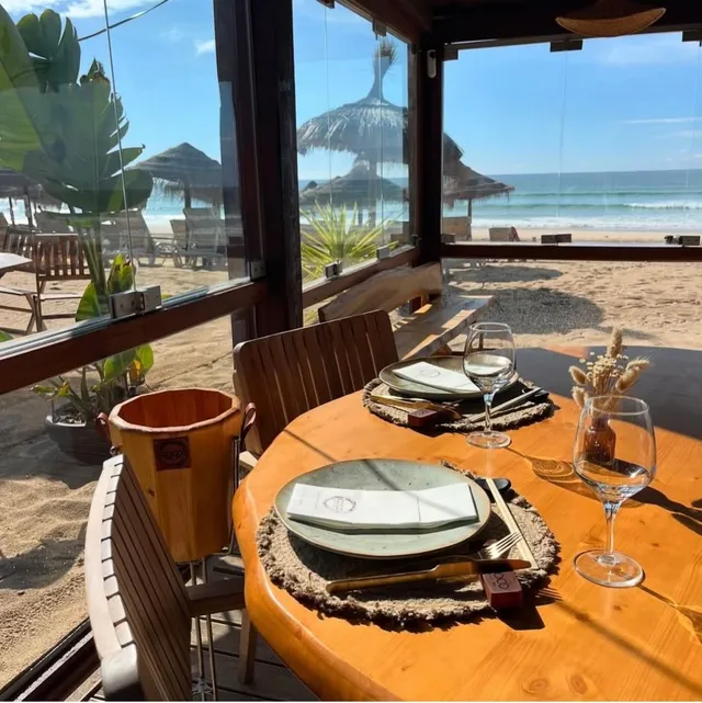 A wooden table set by a glass window facing the sand, straw umbrellas and the ocean waves