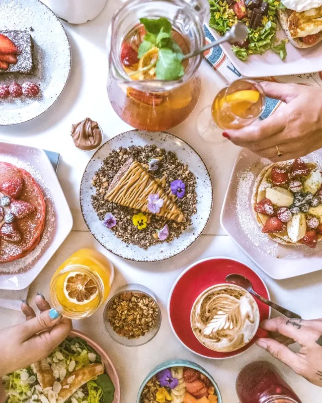 An overhead view of a table loaded with pancakes, cakes, granola, a drinks jug and a cappuccino