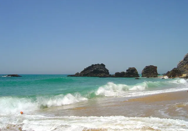 Waves breaking on dark rocks beside a sandy beach and green water