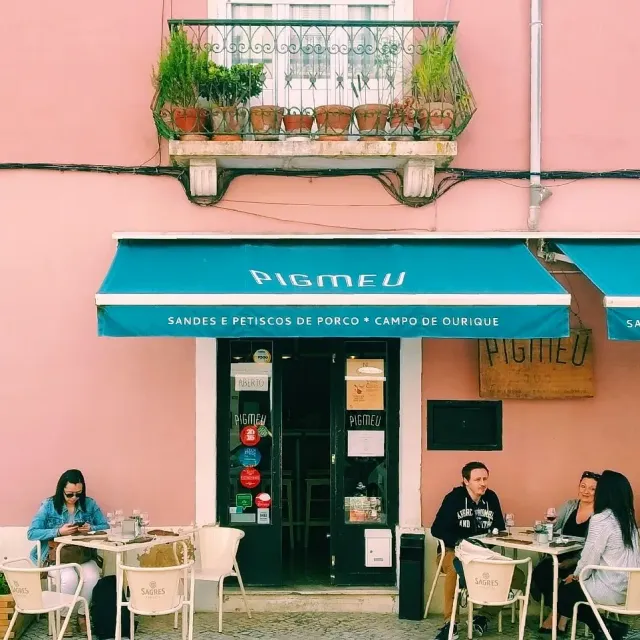 A pink facade with a light blue awning, pots on the balcony and diners at white tables