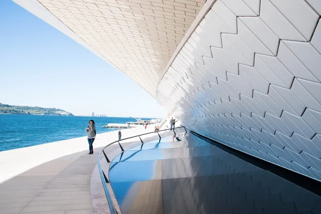 A curving white tiled wall beside a river promenade and blue water