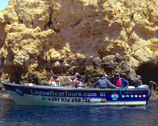 A white tourist boat with passengers hugging a pitted golden rock cliff