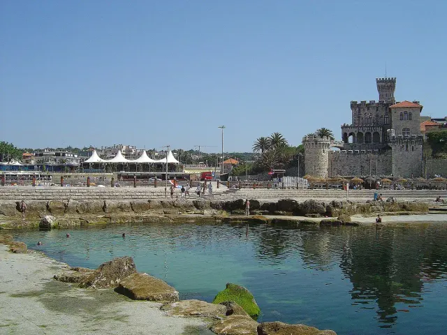 A rocky sea water pool beside a beach with a towered building behind