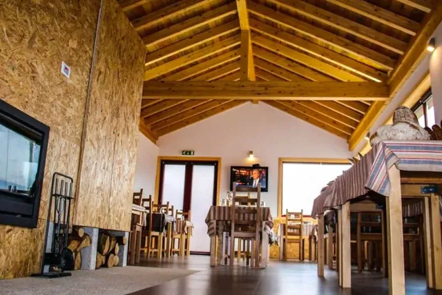 A rustic hall with exposed wooden beams, a wood stove with logs, and tables in striped cloths