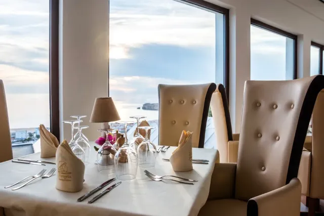 A set table with upturned glasses and tall upholstered chairs facing a window onto the sea