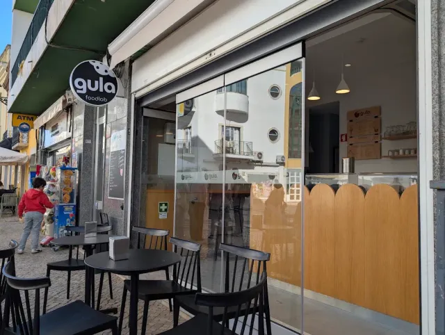 An open shop front with a round sign, black tables and chairs on the sidewalk