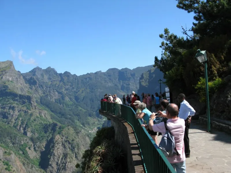 Visitors at the railing of the paved viewing terrace, with the Curral das Freiras valley and its village far below and a jagged ridge across