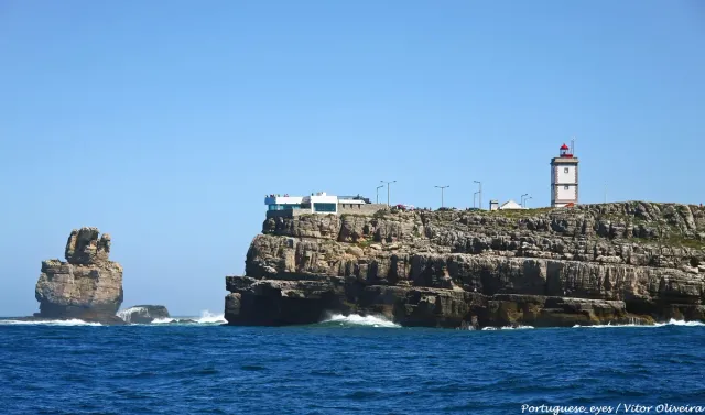 A low lighthouse on a layered cliff above blue sea and an isolated rock