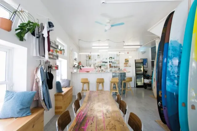 A white space with a long wooden table, colourful surfboards leaning on the wall and a counter with stools