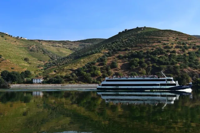 A low cruise boat with passengers on a calm river, hills reflected in the water