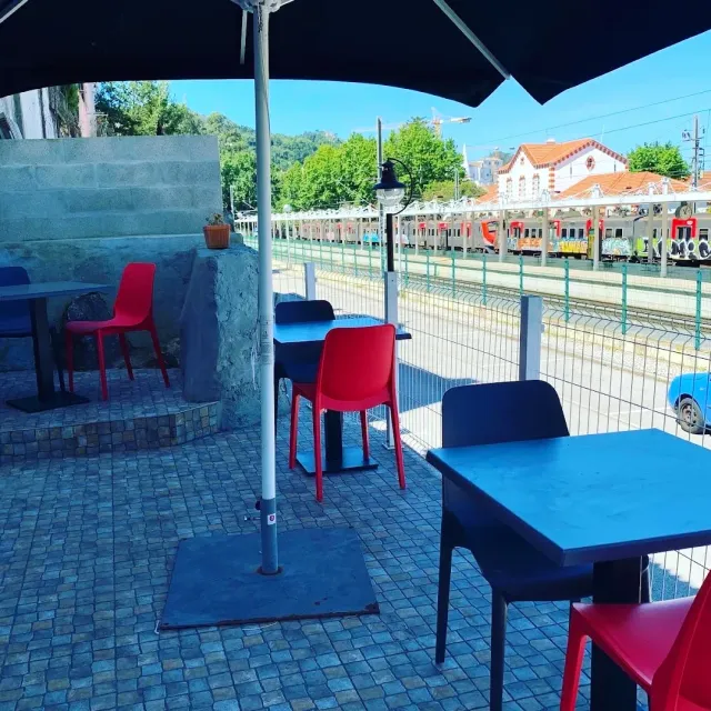 Blue tables and red chairs under a parasol facing a railway line and a station