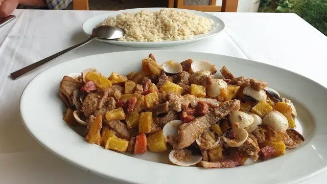 A plate with cubes of meat, potatoes and clams, and a plate of rice behind it