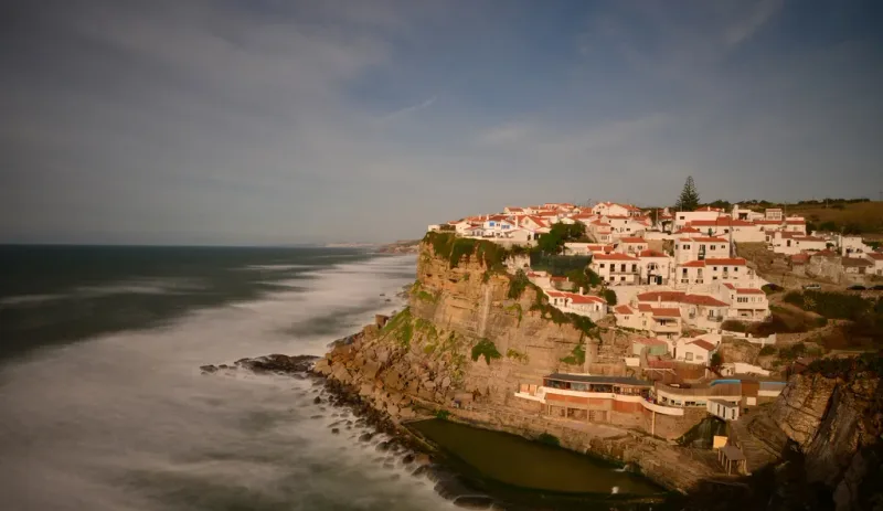 A village of white houses and tiled roofs perched on a clifftop, with waves crashing below