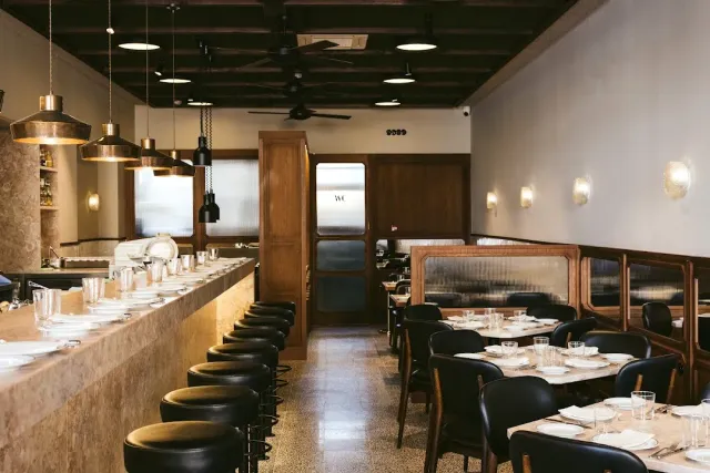 A long counter with black stools, copper lamps and set tables beside it